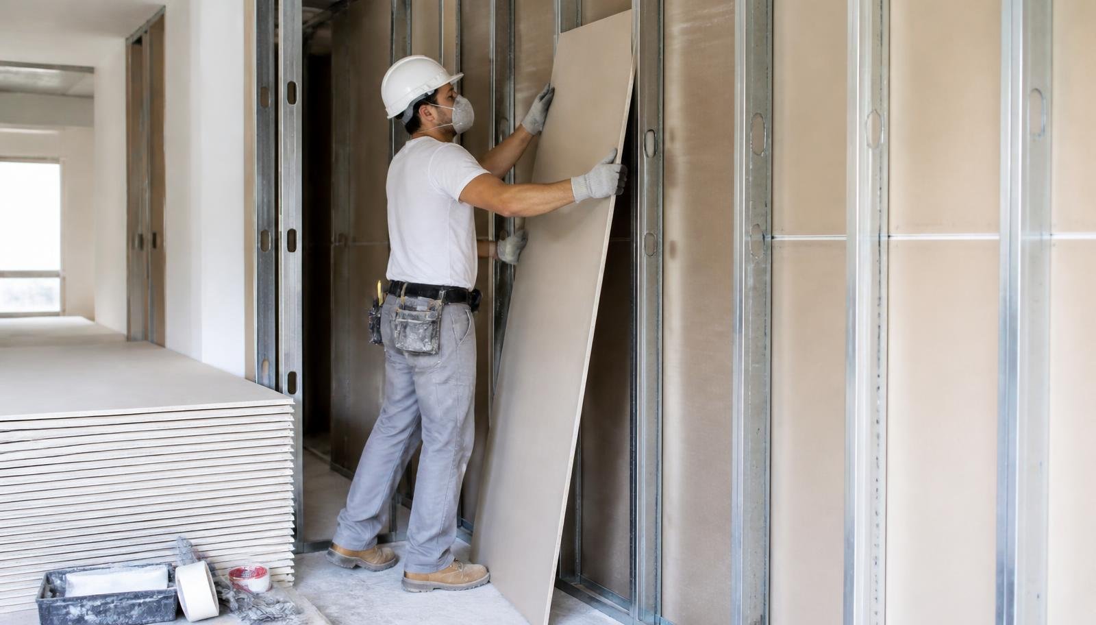 Drywall installer lifting a gypsum board into a metal stud wall frame on an interior job site